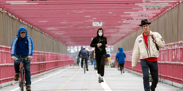 In this Monday, May 11, 2020 photo, a jogger wearing a face masks runs in between a biker and a pedestrian not wearing masks as they make their way over the Williamsburg bridge in New York.New York's governor has ordered masks for anyone out in public who can't stay at least six feet away from other people. Yet, while the rule is clear, New Yorkers have adopted their own interpretation of exactly when masks are required, especially outdoors. (AP Photo/Mary Altaffer)
