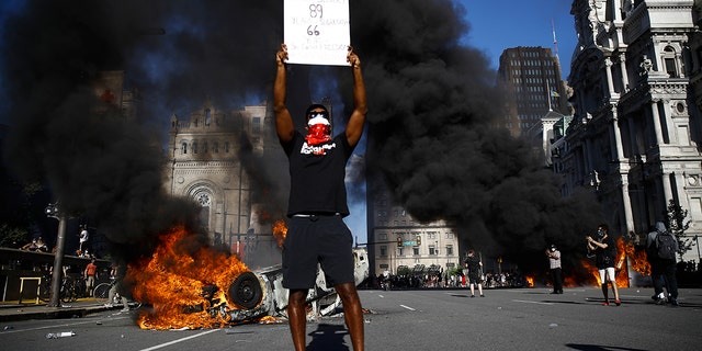 A vehicle is on fire behind a protester holding a sign during a rally on Saturday, May 30, 2020, in Philadelphia, over the death of George Floyd, a black man who died after being taken into police custody in Minneapolis. Floyd died after an officer pressed his knee into his neck for several minutes even after he stopped moving and pleading for air on Memorial Day. (AP Photo/Matt Rourke)