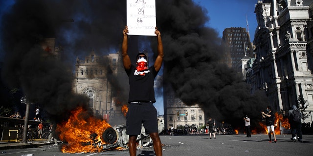 A vehicle is on fire behind a protester holding a sign during a rally on Saturday, May 30, 2020, in Philadelphia, over the death of George Floyd, a black man who died after being taken into police custody in Minneapolis. (Associated Press)