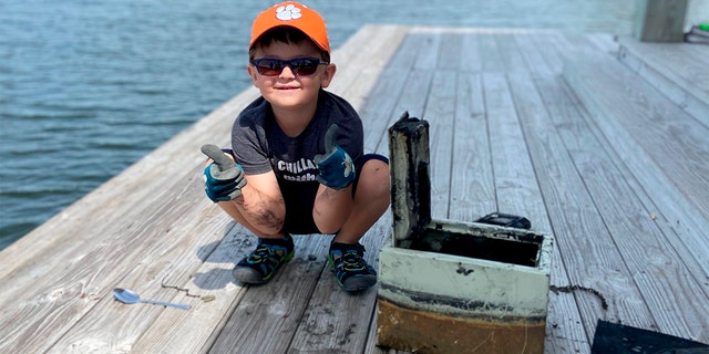 In this May 9, 2020, photo provided by Catherine Brewer, Knox Brewer poses next to a safe he pulled out of Whitney Lake in South Carolina. The 6-year-old was using a magnet attached to a string to fish for metal in the water when he reeled in a lockbox that police said was stolen from a woman who lived nearby eight years ago. (Catherine Brewer via AP)