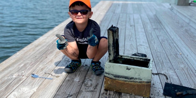 In this May 9, 2020, photo provided by Catherine Brewer, Knox Brewer poses next to a safe he pulled out of Whitney Lake in South Carolina. The 6-year-old was using a magnet attached to a string to fish for metal in the water when he reeled in a lockbox that police said was stolen from a woman who lived nearby eight years ago. (Catherine Brewer via AP)