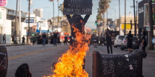 A protester holding a sign stands behind the burning trash cans during a protest over the death of George Floyd, a handcuffed black man in police custody in Minneapolis, in Los Angeles, Saturday, May 30, 2020.