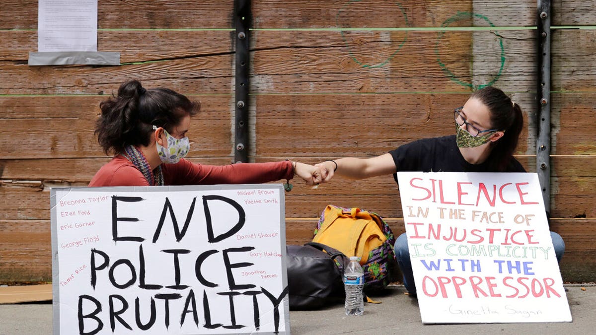 Protesters Kaylee Gore, left, and Amanda Barnes exchange fist bumps as they sit with signs protesting police actions, Thursday, June 4, 2020, in Seattle, following protests over the death of George Floyd in Minneapolis. Seattle's police chief says officers' badge numbers will be prominently displayed following complaints that black bands obscured the digits.