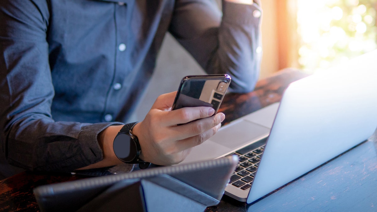 Male hand holding smartphone. Businessman using laptop computer and digital tablet while working in the cafe. Mobile app or internet of things concepts. Modern lifestyle in digital age.