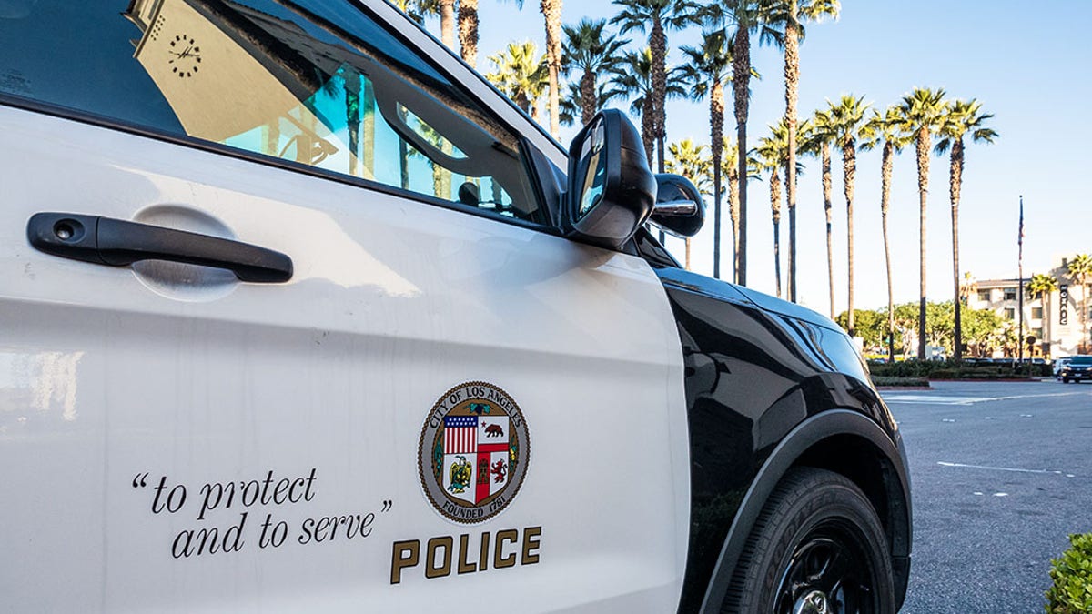 Close-up on the insignia and slogan of a LAPD vehicle, with the reflection of Union Station's tower visible in the car's window.