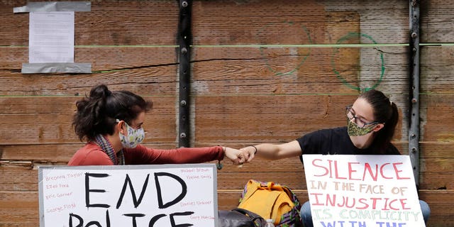 Protesters Kaylee Gore, left, and Amanda Barnes exchange fist bumps as they sit with signs protesting police actions, Thursday, June 4, 2020, in Seattle, following protests over the death of George Floyd in Minneapolis. Seattle's police chief says officers' badge numbers will be prominently displayed following complaints that black bands obscured the digits. 