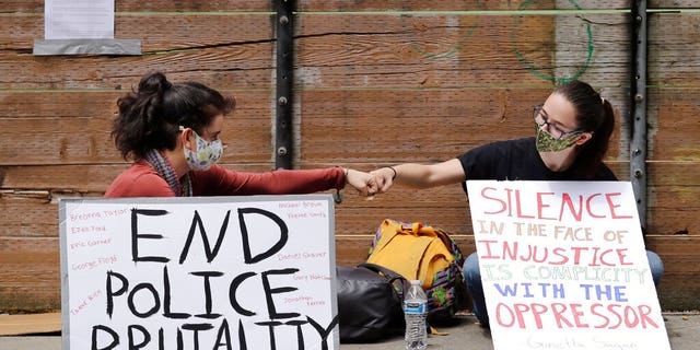 Protesters Kaylee Gore, left, and Amanda Barnes exchange fist bumps as they sit with signs protesting police actions, Thursday, June 4, 2020, in Seattle, following protests over the death of George Floyd in Minneapolis. Seattle's police chief says officers' badge numbers will be prominently displayed following complaints that black bands obscured the digits.
