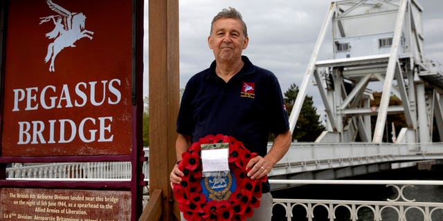 In this photo taken on Friday, June 5, 2020, British expatriate Steven Oldrid holds a poppy wreath as he stands on the site of the original WWII Pegasus Bridge in Benouville, Normandy, France. Due to coronavirus measures many relatives and veterans will not make this years 76th anniversary of D-Day. Oldrid will be bringing it to them virtually as he places wreaths and crosses for families and posts the moments on his facebook page.
