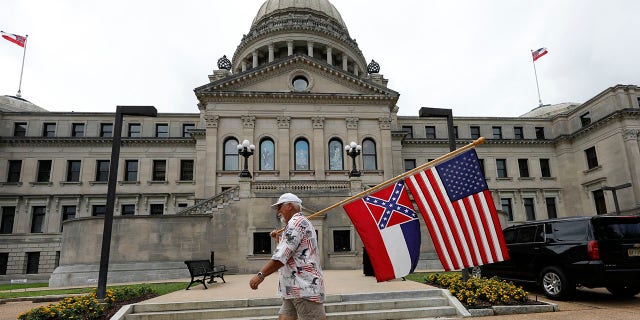 Don Hartness of Ellisville, walks around the Capitol carrying the current Mississippi state flag and the American flag, Saturday, June 27, 2020, in Jackson, Miss. (Associated Press)