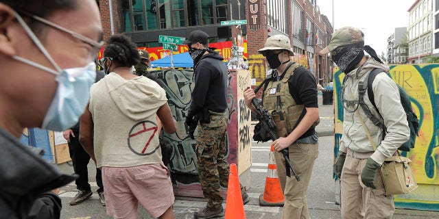 A person who said he goes by the name James Madison, second from right, carries a rifle as he walks Saturday inside what has been named the Capitol Hill Occupied Protest zone in Seattle. Madison is part of the volunteer security team who have been working inside the CHOP zone, and said he and other armed volunteers were patrolling Saturday to keep the area safe. (AP Photo/Ted S. Warren)