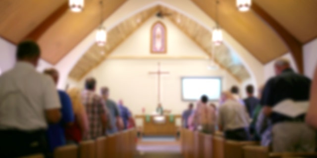 A blurred photo of the inside of a church sanctuary that is filled with people in the pews, and the pastor stands under a large cross at the altar.