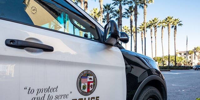 Close-up on the insignia and slogan of a LAPD vehicle, with the reflection of Union Station's tower visible in the car's window.