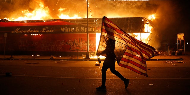 A protester carries the carries a U.S. flag upside, a sign of distress, next to a burning building May 28, in Minneapolis. (AP Photo/Julio Cortez)