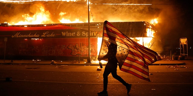 A protester carries the carries a U.S. flag upside, a sign of distress, next to a burning building Thursday, May 28, 2020, in Minneapolis. Protests over the death of George Floyd, a black man who died in police custody Monday, broke out in Minneapolis for a third straight night. (AP Photo/Julio Cortez)
