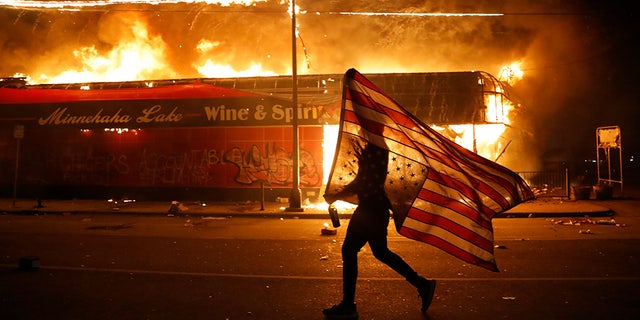 A protester carries the carries a U.S. flag upside, a sign of distress, next to a burning building May 28, in Minneapolis. (AP Photo/Julio Cortez)