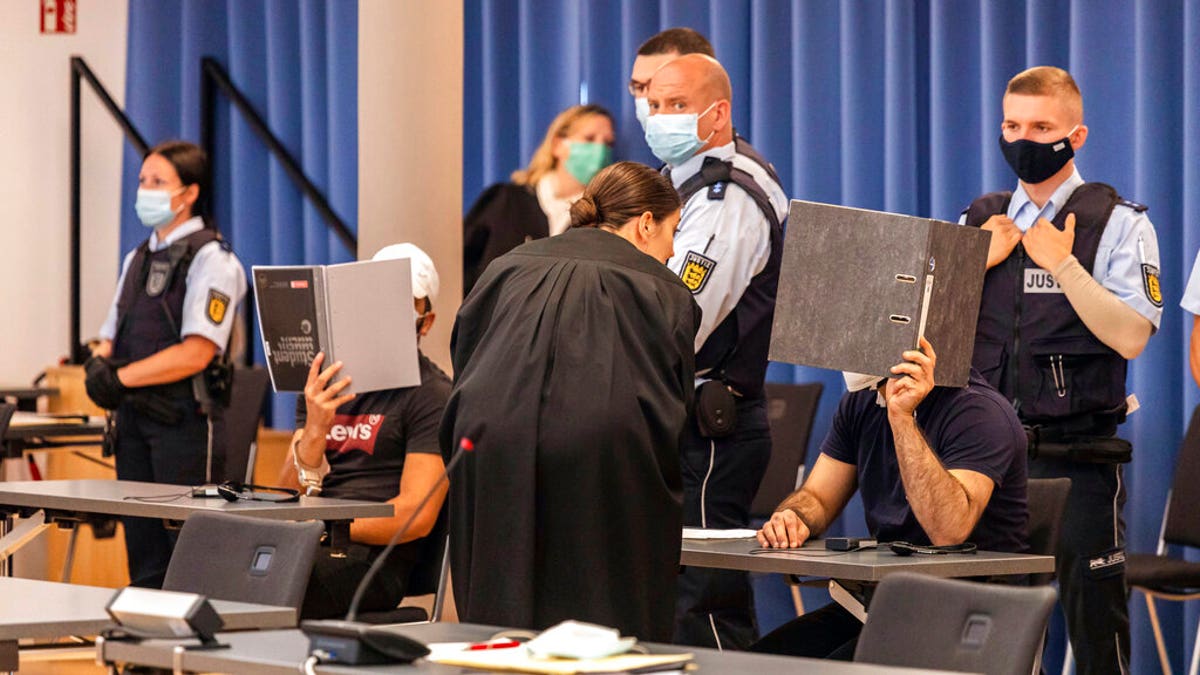 Two defendants are holding folders in front of their faces as a screen, while a defense attorney is talking to the right defendant and judicial officers are standing behind the defendants at a trial in Freiburg, Germany. 