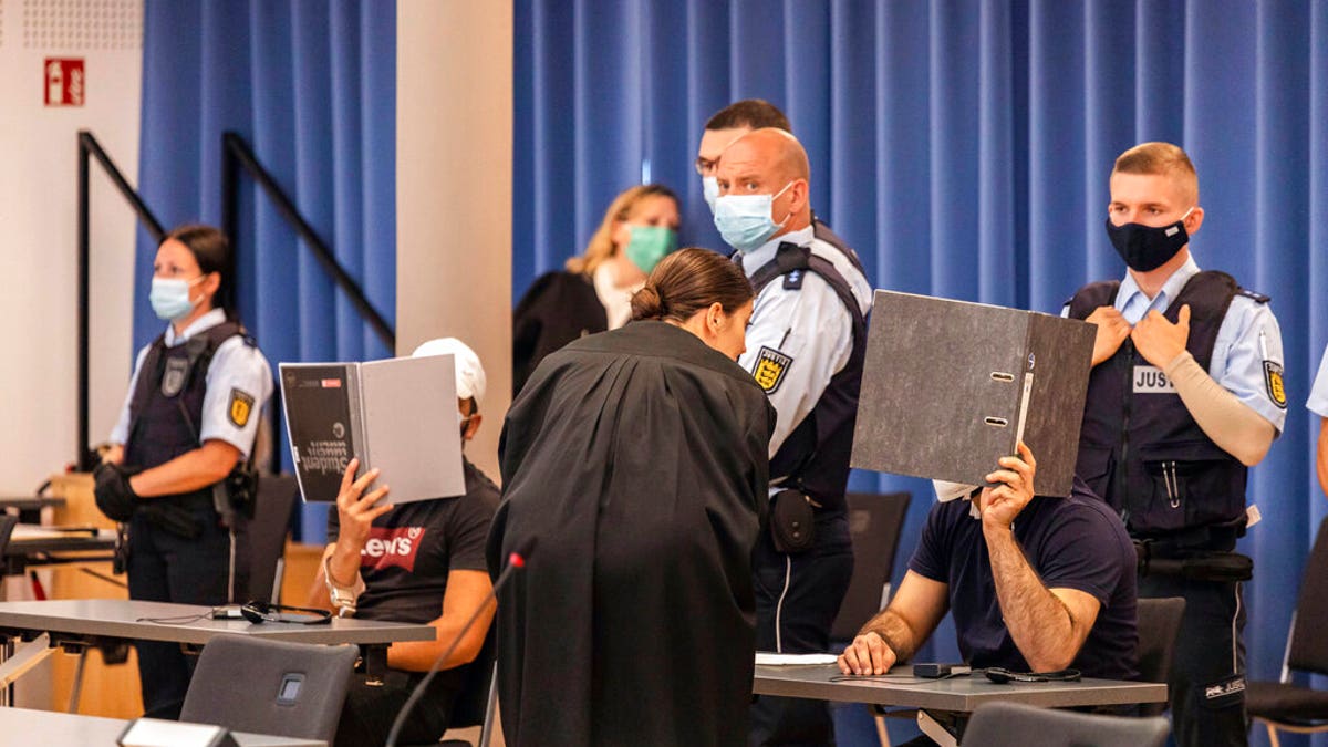 Two defendants are holding folders in front of their faces as a screen, while a defense attorney is talking to the right defendant and judicial officers are standing behind the defendants at a trial in Freiburg, Germany.