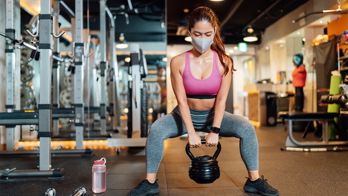 A female athlete is wearing a protective face mask and lifting a kettlebell in a gym. (Stock image)