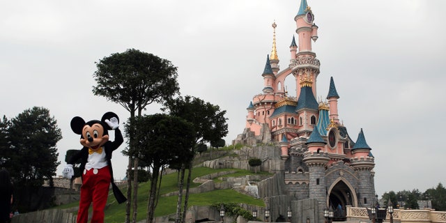 In this June 8, 2018, file photo, a person dressed as Mickey Mouse poses in front of the castle of Sleeping Beauty at Disneyland Paris, in Chessy, France.