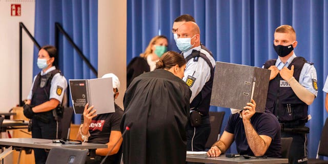 Two defendants are holding folders in front of their faces as a screen, while a defense attorney is talking to the right defendant and judicial officers are standing behind the defendants at a trial in Freiburg, Germany.