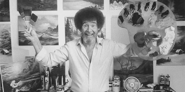 TV painting instructor/artist Bob Ross jubilantly holding up paint pallette & brushes as he stands in front of a wall covered with his landscape paintings in his studio.