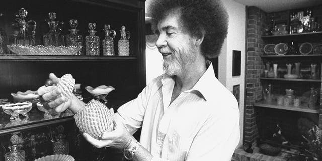 TV painting instructor/artist Bob Ross examining a piece from his collection of turn-of-the-century Victorian opalescent glass next to a cabinet full of decanters & dishes at home.