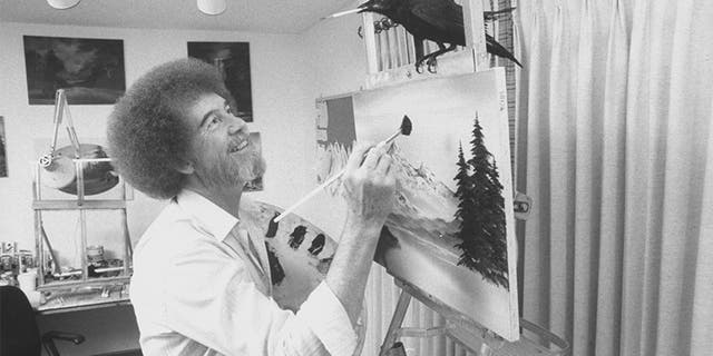 TV painting instructor/artist Bob Ross painting one of his landscapes as his pet crow holds a paintbrush in its beak while perched on top of an easel in his studio home.