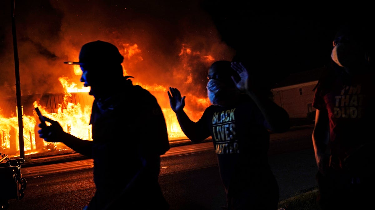 Protesters walk past police with their arms up, late Monday, Aug. 24, 2020, in Kenosha, Wis., as a building burns in the background. Protests have erupted following the police shooting of Jacob Blake a day earlier. (AP Photo/David Goldman)