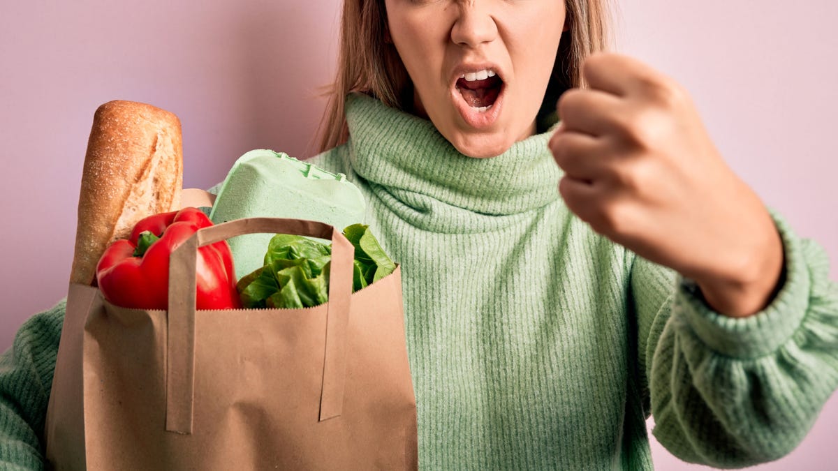 Young beautiful blonde girl holding fresh groceries paper bag over pink isolated background annoyed and frustrated shouting with anger, crazy and yelling with raised hand, anger concept
