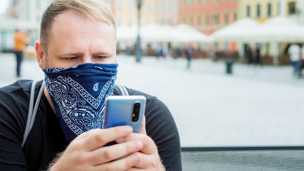 Adult man in a bandana as a face mask uses a smartphone in the city cafe terrace.