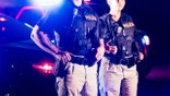 Cropped view of two multi-ethnic police officers standing in front of patrol cars, wearing bulletproof vests and duty belts. The policewoman is a mature African-American woman in her 40s. Her partner is a mid adult man in his 30s. It is nighttime and the emergency lights on top of the vehicles are flashing.