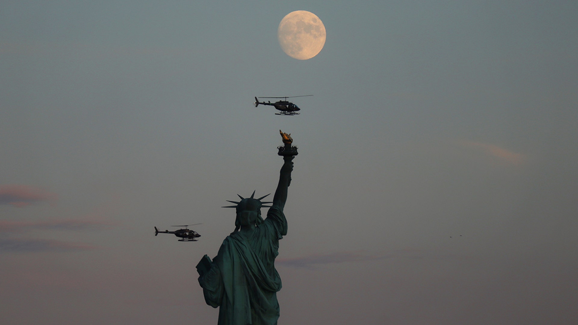 The moon rises behind the Statue of Liberty as two helicopters fly past in New York City, Aug. 1, 2020