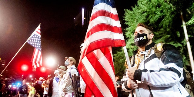 Navy veteran Adam Winther holds a flag while forming a "Wall of Vets" during a Black Lives Matter protest at the Mark O. Hatfield United States Courthouse on Friday, July 31, 2020, in Portland, Ore. Following an agreement between Democratic Gov. Kate Brown and the Trump administration to reduce federal officers in the city, nightly protests remained largely peaceful without major confrontations between demonstrators and officers. (AP Photo/Noah Berger)