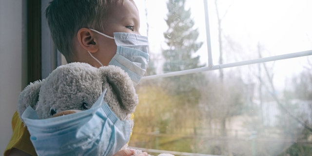 Boy and his teddy bear both in protective medical masks sits on windowsill and looks out window. 