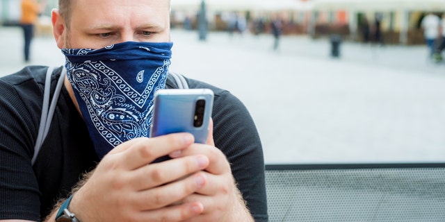 Adult man in a bandana as a face mask uses a smartphone in the city cafe terrace. 