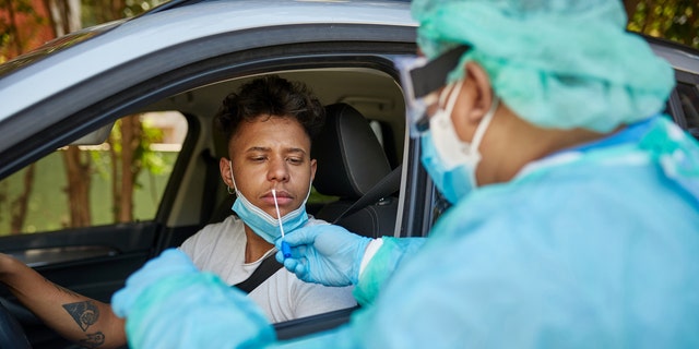 一名身穿防护工作服的医疗保健专业人员准备对一名 20 岁出头的非洲裔加勒比男子在驾驶位置进行鼻拭子测试。