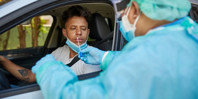 Healthcare professional in protective workwear preparing to do nasal swab testing on Afro-Caribbean man in early 20s at drive-thru site.