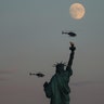 The moon rises behind the Statue of Liberty as two helicopters fly past in New York City, Aug. 1, 2020