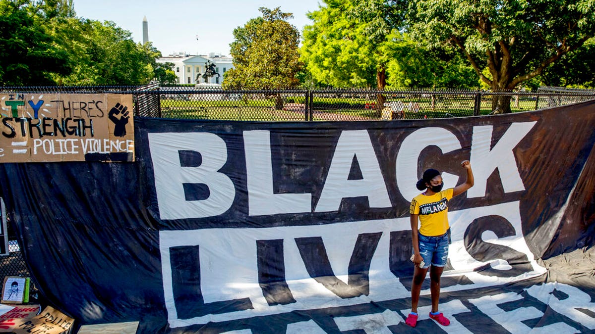 The White House is visible behind a woman who holds her fist up as she poses for a photograph with a large banner that reads Black Lives Matter hanging on a security fence in Washington, after days of protests over the death of George Floyd. June 8, 2020. (AP Photo/Andrew Harnik, File)