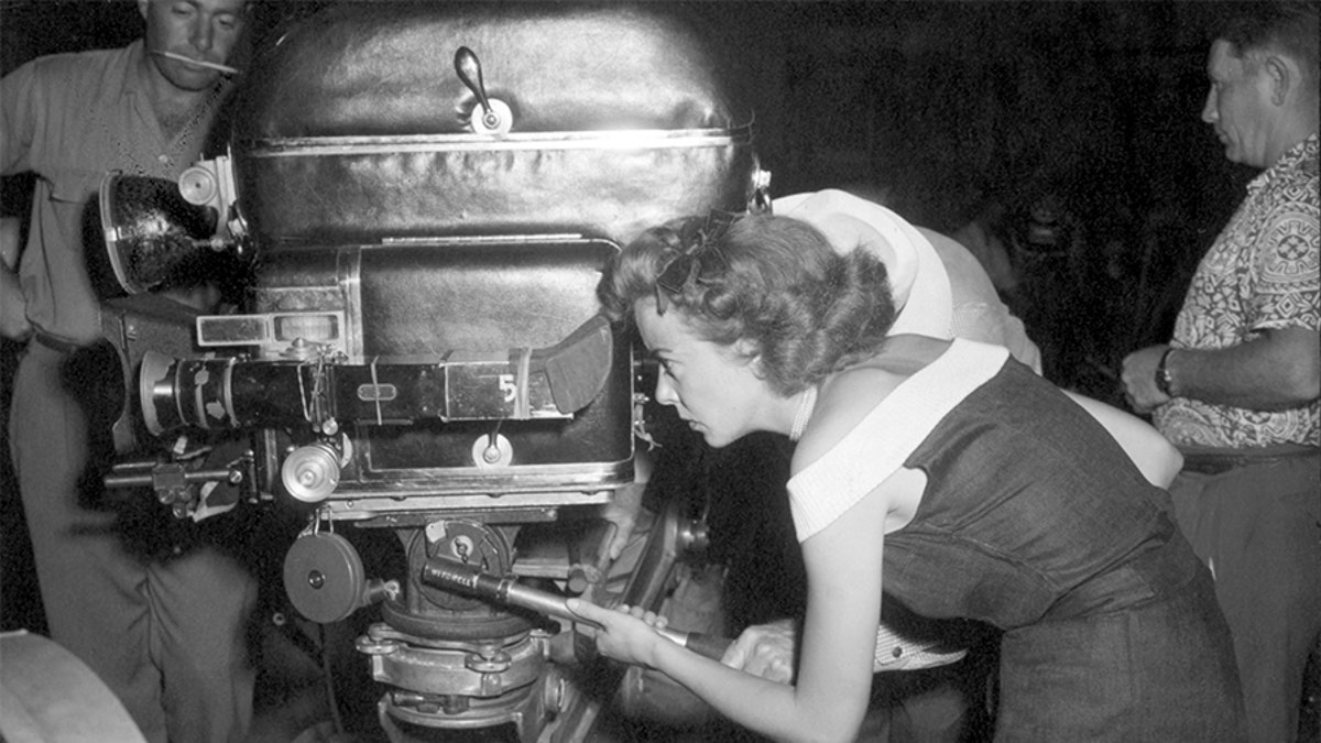Ida Lupino directs one of the scenes from her film 'Mother of a Champion.' She is shown peering through the movie camera. Undated photograph.