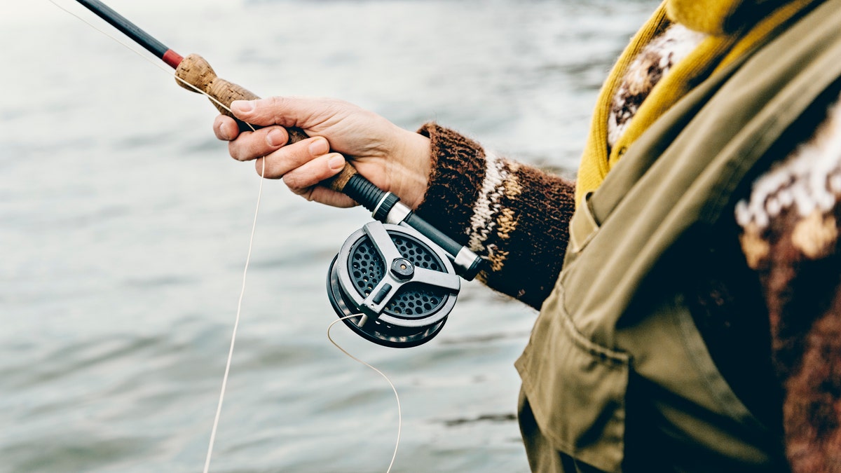 Close up portrait of a fly fisherman's hands as she works the line and reel. She is wearing a green cotton gillet,waders, a patterned sweater and a mustard coloured scarf. Colour, horizontal format photographed against a calm sea with lots of copy space. Photographed on location at Nordfeld Strand on the island of Møn in Denmark.