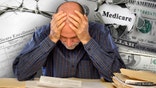 A senior adult man sitting at a table or desk stacked with papers and envelopes looking down with his hands on his head.