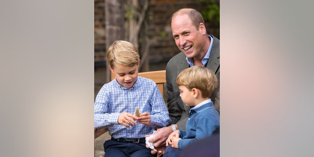 Prince William, Duke of Cambridge and Prince Louis of Cambridge watch as Prince George of Cambridge holds the tooth of a giant shark given to him by Sir David Attenborough in the gardens of Kensington Palace