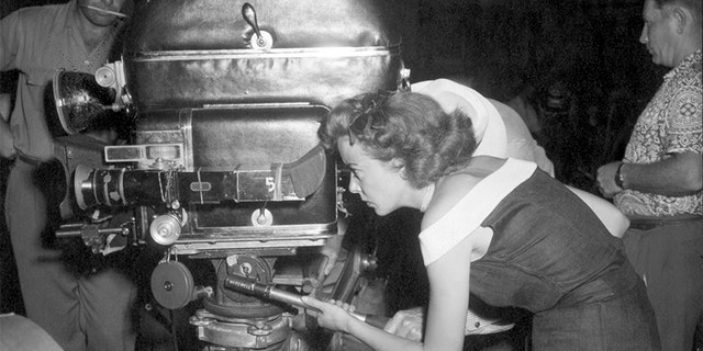 Ida Lupino directs one of the scenes from her film 'Mother of a Champion.' She is shown peering through the movie camera. Undated photograph.