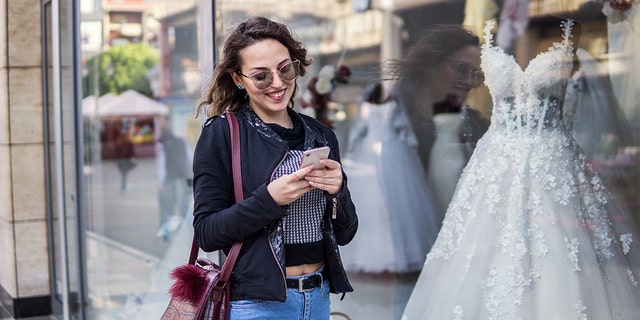 Woman using a mobile phone in the city, outdoor portrait of cheerful young woman texting on a smart cell phone in front of a clothing store,  wedding dresses in the background. File photo.