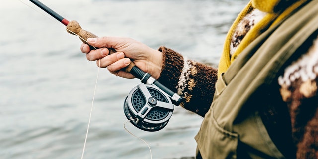 Close up portrait of a fly fisherman's hands as she works the line and reel. She is wearing a green cotton gillet,waders, a patterned sweater and a mustard coloured scarf. Colour, horizontal format photographed against a calm sea with lots of copy space. Photographed on location at Nordfeld Strand on the island of Møn in Denmark.