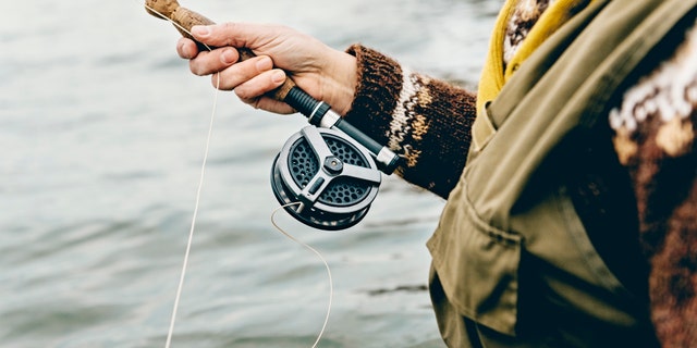 Close up portrait of a fly fisherman's hands as she works the line and reel. She is wearing a green cotton gillet,waders, a patterned sweater and a mustard coloured scarf. Colour, horizontal format photographed against a calm sea with lots of copy space. Photographed on location at Nordfeld Strand on the island of Møn in Denmark.