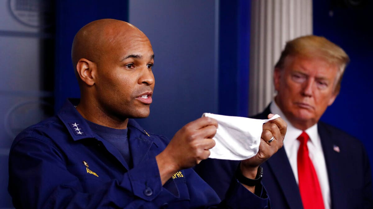 In this April 22, photo President Trump watches as U.S. Surgeon General Jerome Adams holds up his face mask at a coronavirus briefing in the White House. Adams was cited for being in a closed Hawaii park in August while in the islands helping with surge testing amid a spike in coronavirus cases. (AP Photo/Alex Brandon, File)