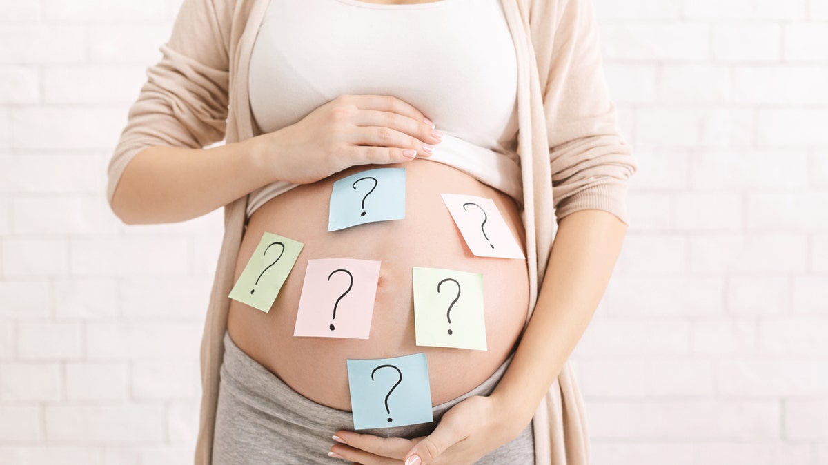 Woman holds pregnant belly with question mark sticky notes