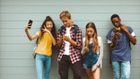 College friends standing outdoors looking using their cell phones. Young girl taking a selfie standing outdoors against a wall with her friends looking at their mobile phones. (iStock)
